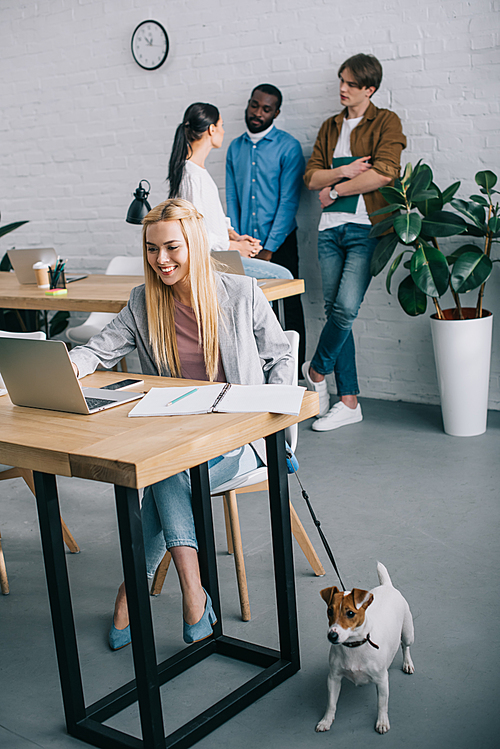 smiling businesswoman using laptop and holding jack russell terrier on leash and coworkers having meeting behind in modern office