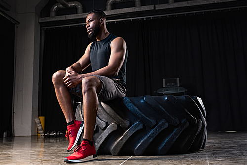 low angle view of pensive african american sportsman sitting on tyre and looking away