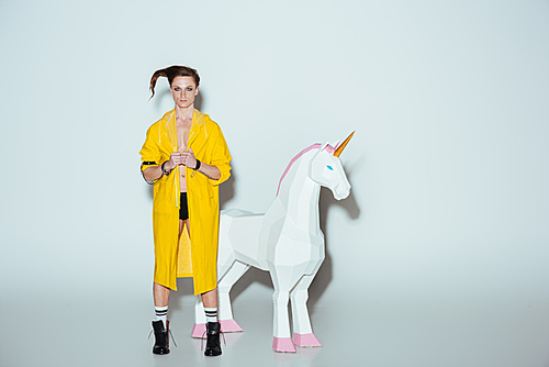 fashionable man with hairstyle in yellow raincoat standing with big unicorn toy, on grey