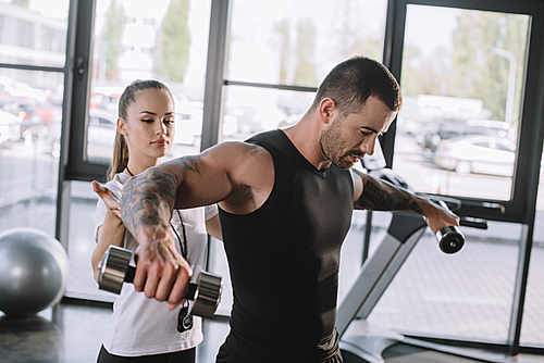 female personal trainer helping sportsman to do exercises with dumbbells at gym