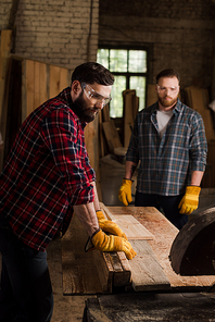 bearded carpenter in safety googles using machine saw and partner standing behind at sawmill