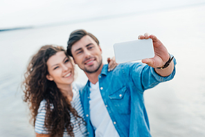 selective focus of smiling couple taking selfie on smartphone near sea