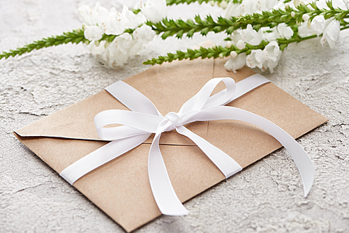 beige envelope with white ribbon near flowers on grey textured surface