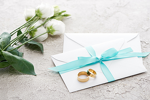 golden rings on white envelopes with ribbon near eustoma flowers on textured surface