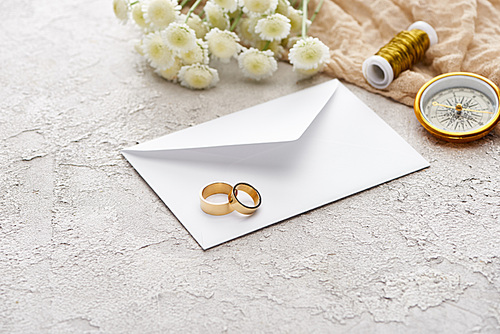 wedding rings on white envelope near chrysanthemums, beige sackcloth, spool and golden compass on textured surface