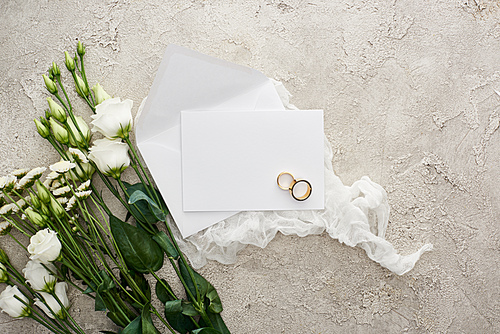 top view of wedding rings on empty card on white cheesecloth near flowers on textured surface