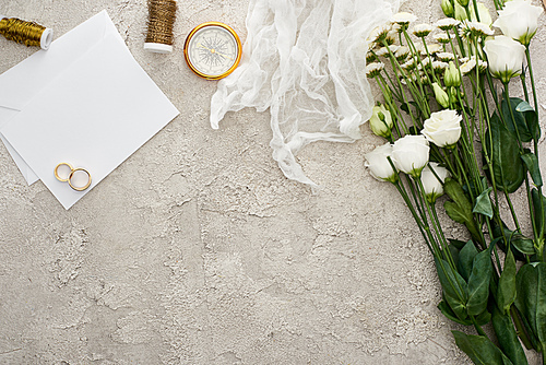 top view of wedding rings on blank invitation card, white cheesecloth and flowers on textured surface