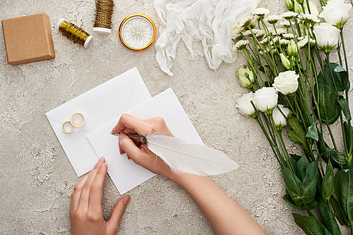 cropped view of woman writing on empty card near wedding rings, compass, cheesecloth, flowers and gift box on textured surface