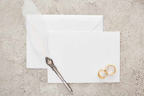 top view of wedding rings and quill pen on white empty card on textured surface