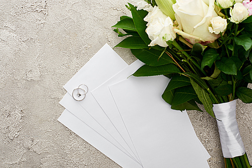 top view of silver rings on invitation cards near bouquet on textured surface