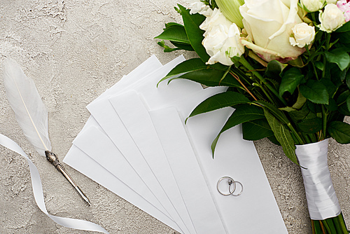 top view of silver rings on envelopes near bouquet, white ribbon and quill pen on grey textured surface