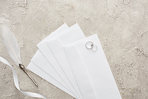 top view of silver rings on envelopes near white ribbon and quill pen on grey textured surface