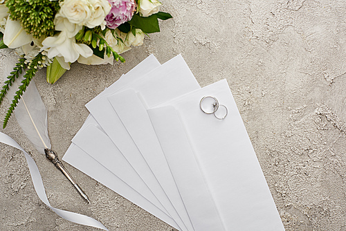 top view of wedding rings on envelopes near quill pen, white ribbon and bouquet on textured surface