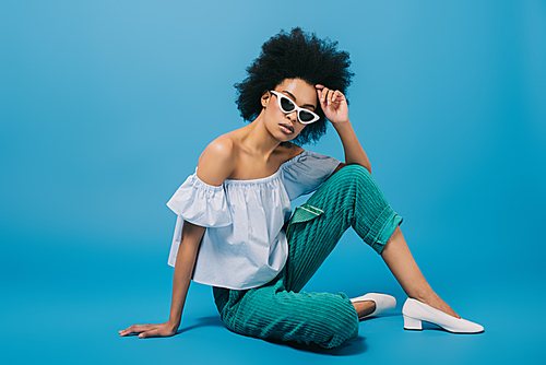attractive young woman in stylish off-the-shoulder top and sunglasses sitting on floor on blue