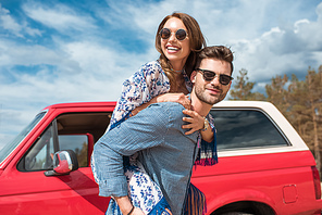 young smiling couple in sunglasses piggybacking near red car