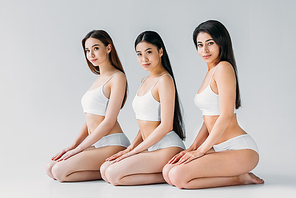 young charming multicultural women posing in white underwear, isolated on grey