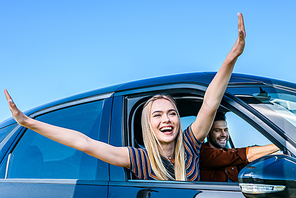 happy young woman with wide arms leaning out from car window while her boyfriend driving car