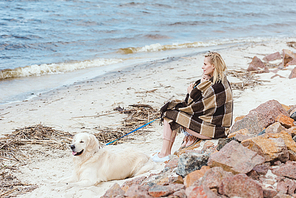 blonde woman wrapped in blanket sitting on sea shore with dog