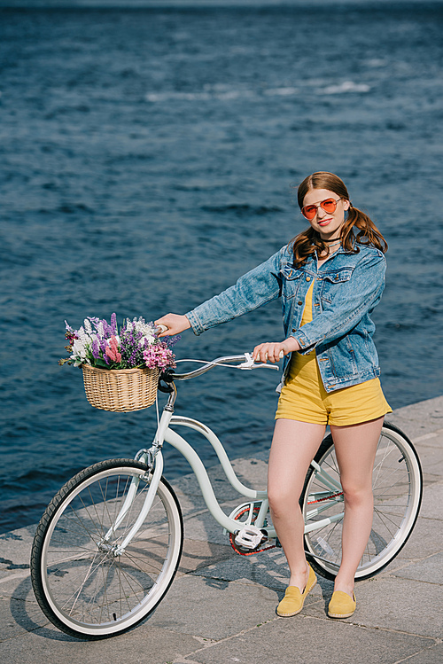 high angle view of girl in sunglasses smiling at camera while standing with bicycle near river