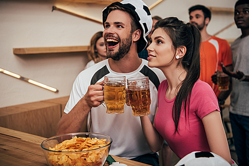 excited man in soccer ball hat clinking beer glasses with girlfriend while their multicultural friends watching football match at bar