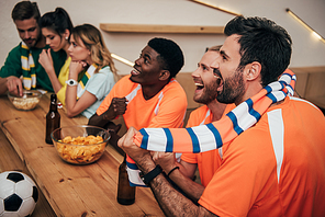 side view of happy young multicultural friends in orange fan t-shirts celebrating goal in soccer match while their friends sitting upset on other side of bar counter