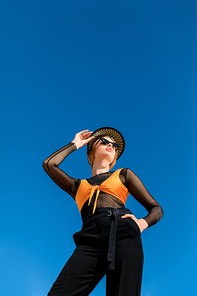 bottom view of girl posing in trendy sunglasses and stylish hat, blue sky on background
