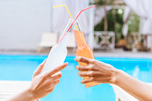 close-up partial view of young women clinking bottles of summer drinks while resting at poolside