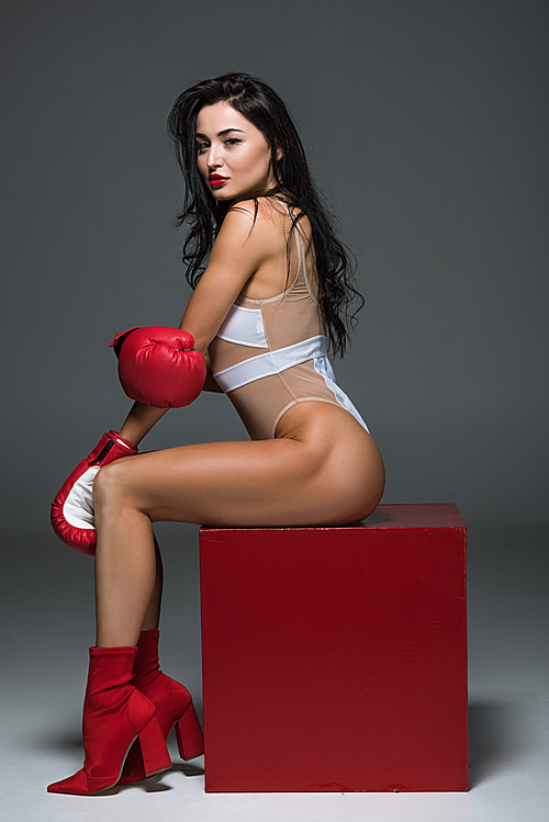 side view of sexy sportive woman in white leotard and boxing gloves sitting on red cube on grey
