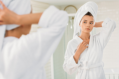 beautiful young woman in bathrobe and towel on head looking at mirror in bathroom