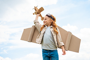 low angle view of child in pilot costume holding wooden toy plane against blue cloudy sky