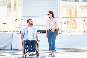 handsome boyfriend in wheelchair and girlfriend walking and  looking at each other on street