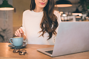 partial view of female freelancer sitting with laptop and coffee cup at table in cafe