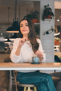 beautiful young woman looking away and holding eyeglasses while sitting at table with coffee cup in cafe