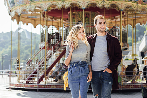 heterosexual couple in autumn outfit walking and cuddling near carousel in amusement park