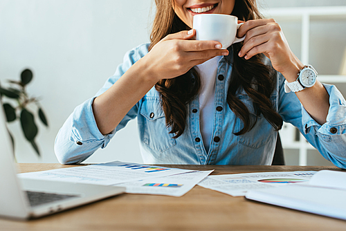 cropped shot of smiling businesswoman with cup of coffee at workplace with papers and laptop in office