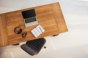 top view of laptop with blank screen, headphones and notebook on wooden table with office chair near by