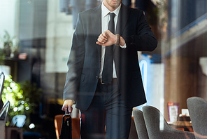 partial view of businessman with suitcase checking time while walking in cafe
