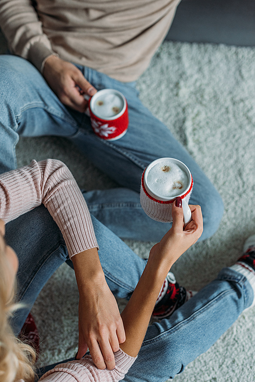 cropped image of couple sitting on carpet and holding cups of cappuccino at home, christmas concept
