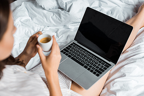 partial view of woman with coffee using laptop with blank screen in bed