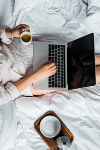 cropped view of woman with coffee cup using laptop in bed