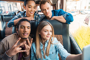happy multiethnic friends taking selfie on smartphone during trip on travel bus