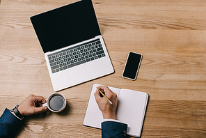 partial view of businessman writing in notebook at workplace with coffee and gadgets