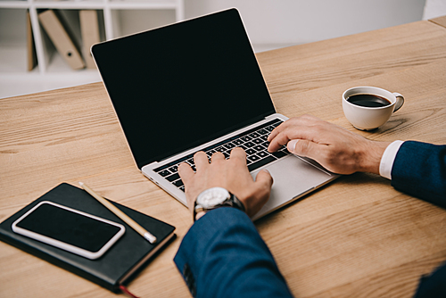 cropped view of businessman typing on laptop at workplace with smartphone and cup of coffee