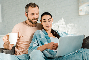 husband holding cup of coffee while beautiful wife using laptop on couch