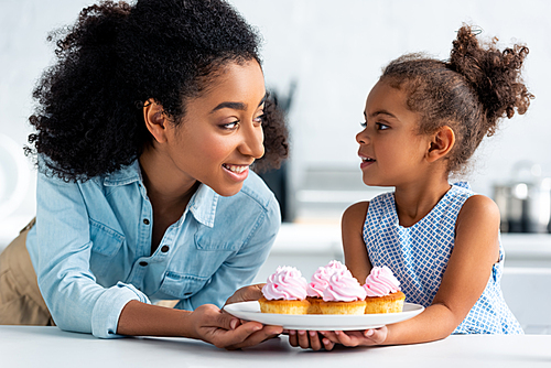 smiling african american mother and daughter holding homemade cupcakes and looking at each other in kitchen