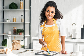 beautiful smiling african american girl in apron applying oil on unbaked cookies in kitchen and 