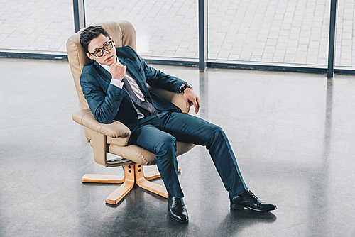 high angle view of confident asian businessman in eyeglasses sitting with hand on chin in office