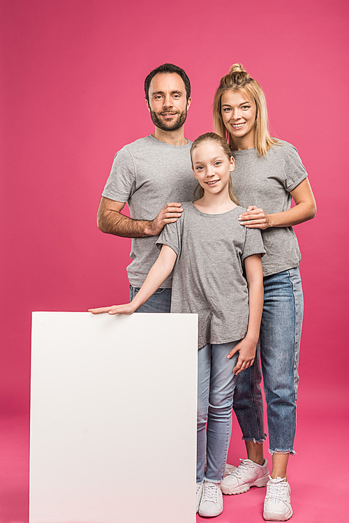 smiling parents and daughter posing with blank placard, isolated on pink