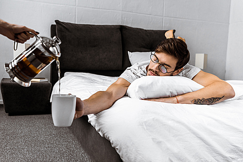 cropped shot of someone pouring tea from teapot while sleepy man in eyeglasses lying on bed and holding cup