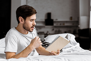handsome young man holding eyeglasses and reading book while lying on bed